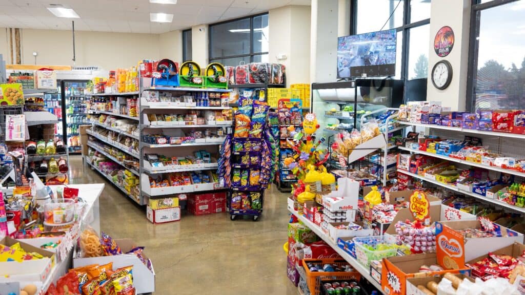 Interior of a convenience store with shelves stocked with snacks, drinks, and candy; security monitors and a clock are visible on the wall by large windows.