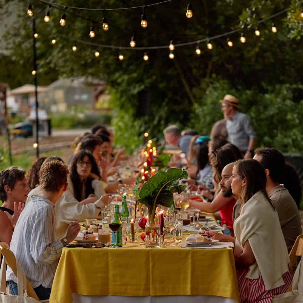 A group of people sit at a long outdoor table decorated with lights in Centennial Park, eating and talking during a gathering inspired by French fashion.
