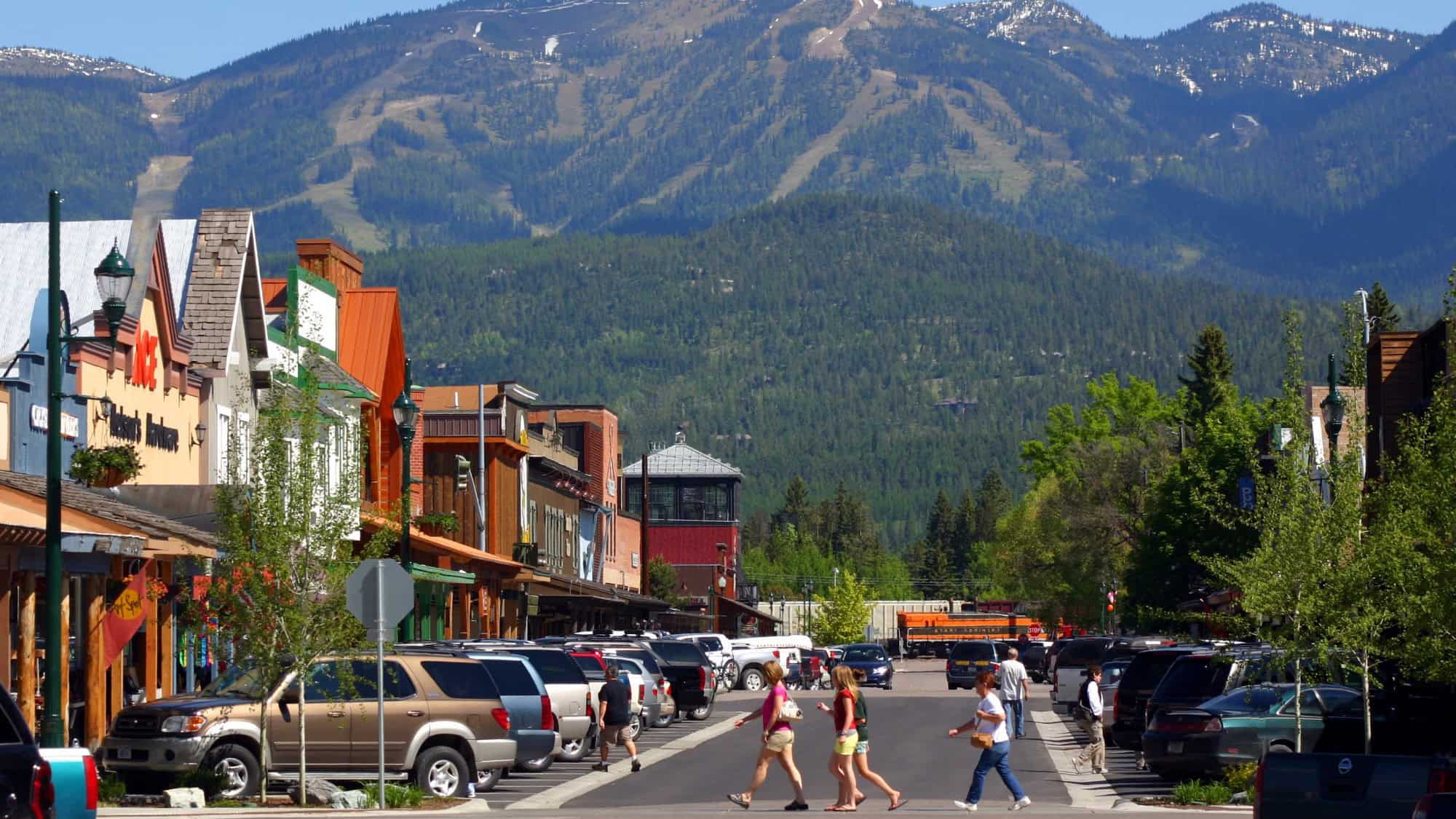 People cross a street lined with parked cars and shops in a small town, with a forested mountain visible in the background under a partly cloudy sky.