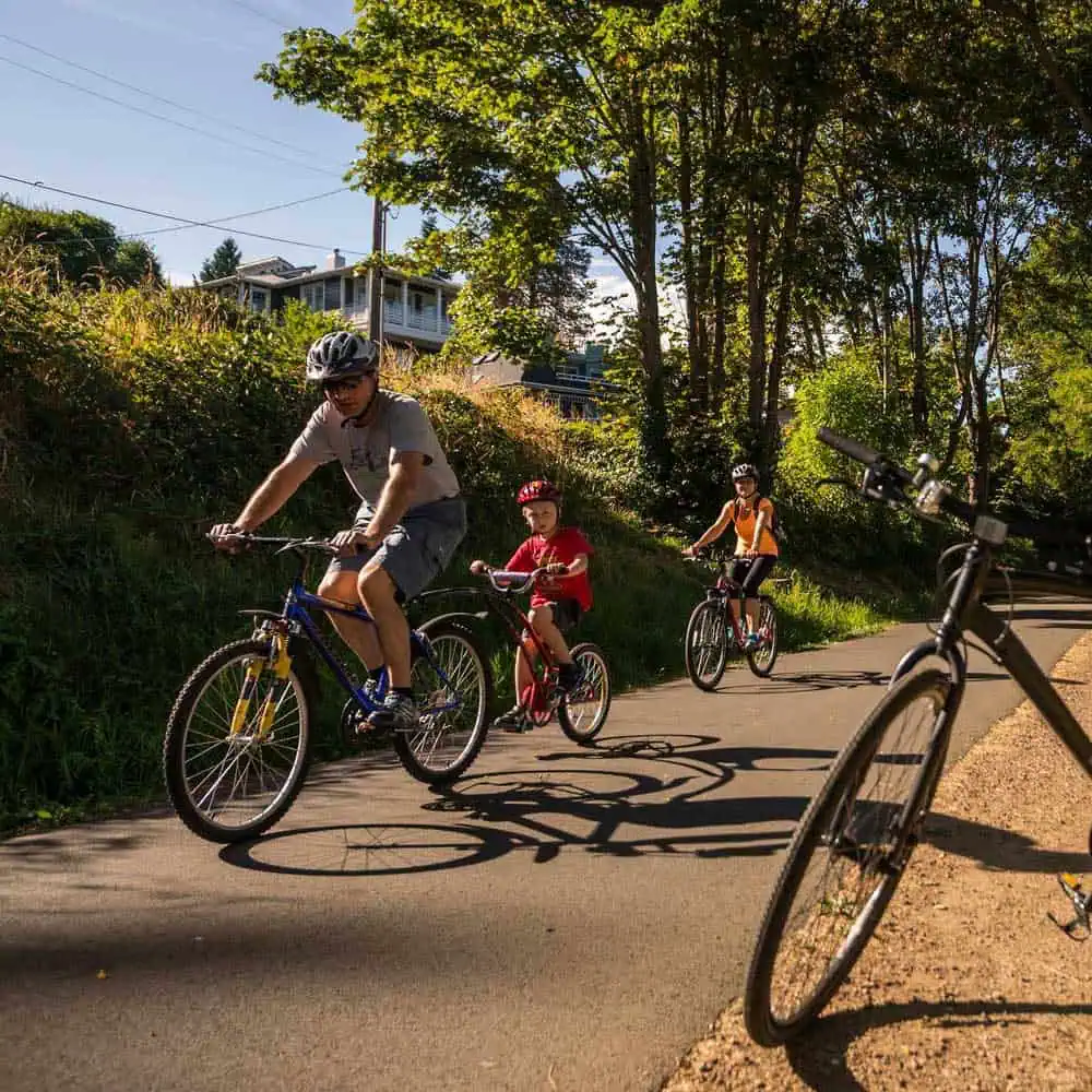 Three people wearing helmets ride bicycles on a paved path surrounded by trees on a sunny day—a perfect Father’s Day outing; another bicycle is parked nearby in the foreground.