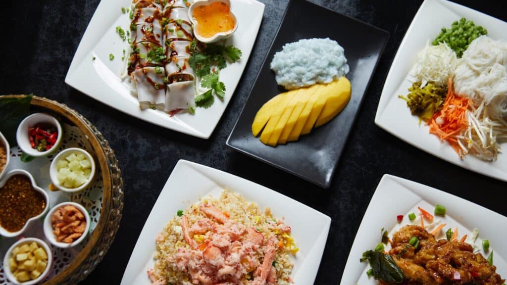 Five plates of Thai food, including mango sticky rice, steamed rolls, fried rice, stir-fried dish, and assorted condiments, displayed on a dark surface.