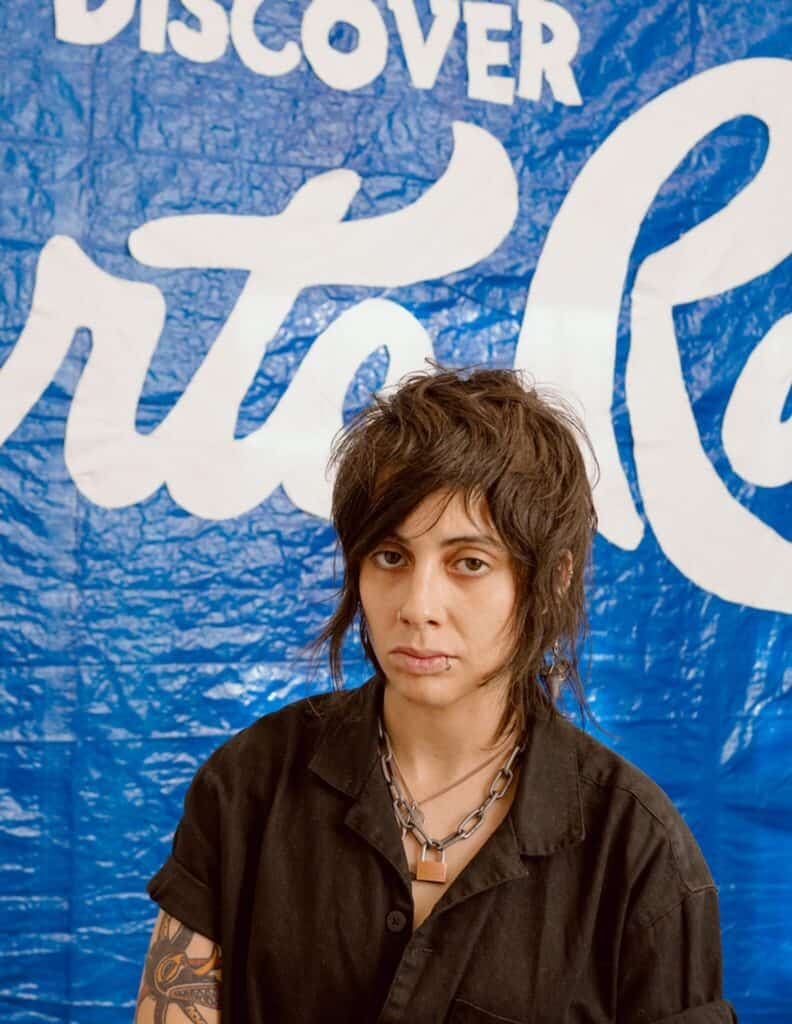 A person with shaggy dark hair and tattoos, wearing a black shirt and layered necklaces, stands in front of a blue and white sign that reads “DISCOVER.”.
