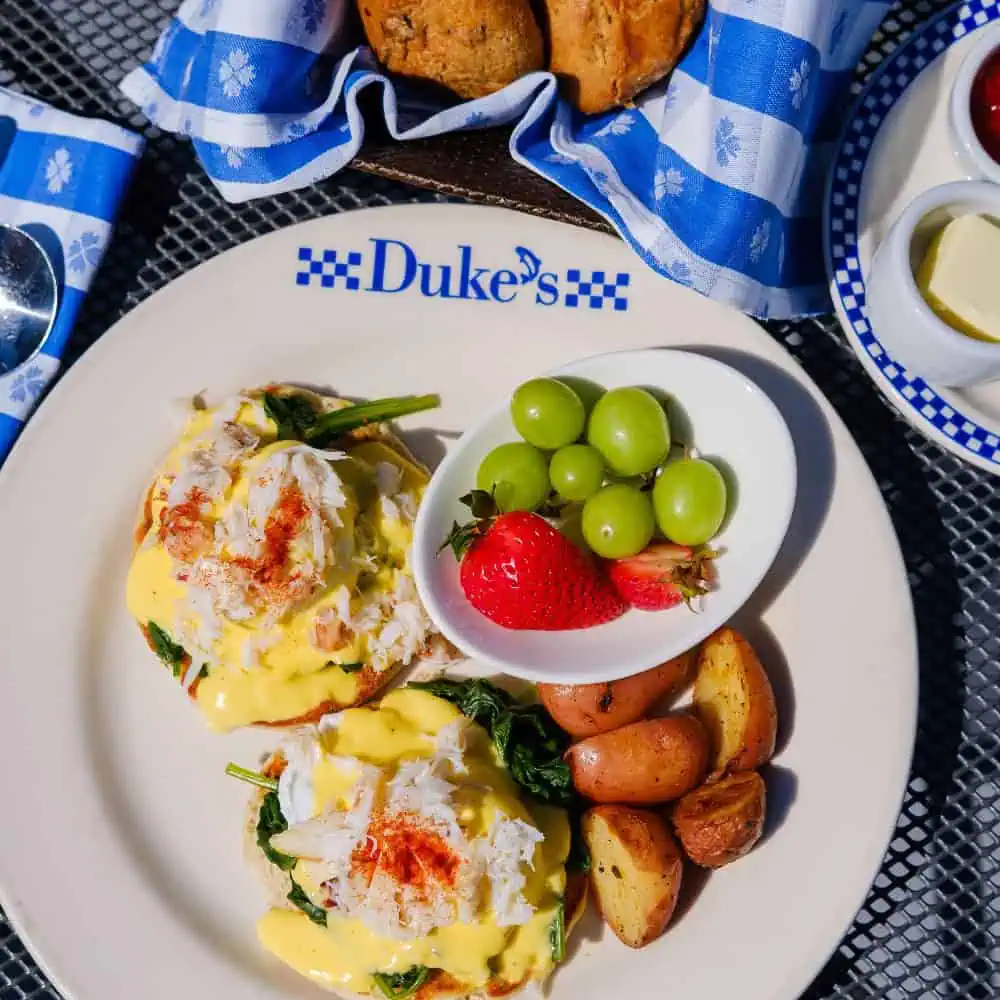 A plate of eggs benedict with crab, roasted potatoes, and a side of green grapes and a strawberry, served at Duke's restaurant—a delicious Father’s Day brunch idea to consider for your Gift Guide. Cornbread and condiments are in the background.