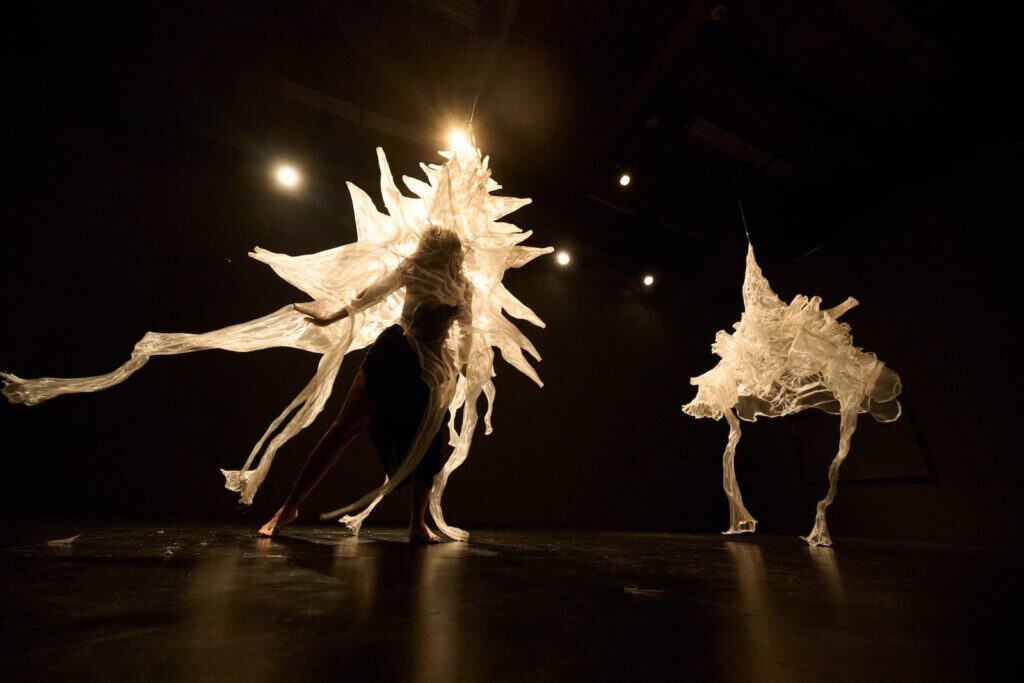 Two figures on a dark stage wear abstract costumes made of translucent, spiky material, illuminated by spotlights from above.