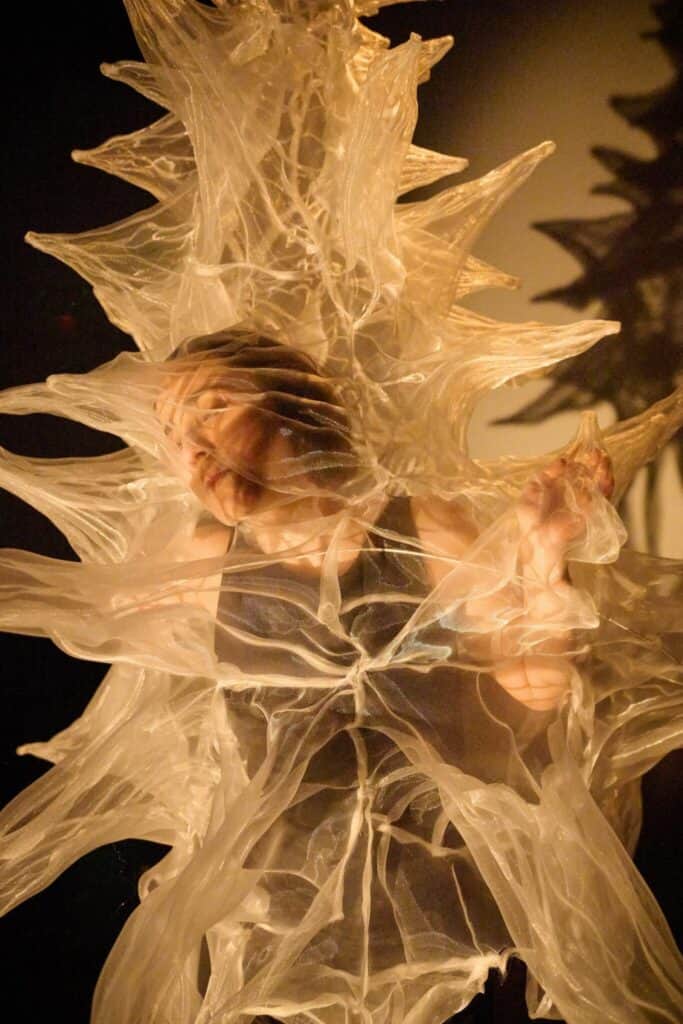 A person presses against translucent, spiky plastic material, creating distorted shapes and shadows under dramatic lighting.