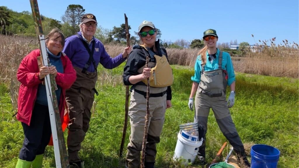 Four people stand outdoors in a grassy area near a trailhead, wearing waders and outdoor gear, holding tools and buckets, with tall grass and blue sky providing scenic Trailhead Direct views in the background.