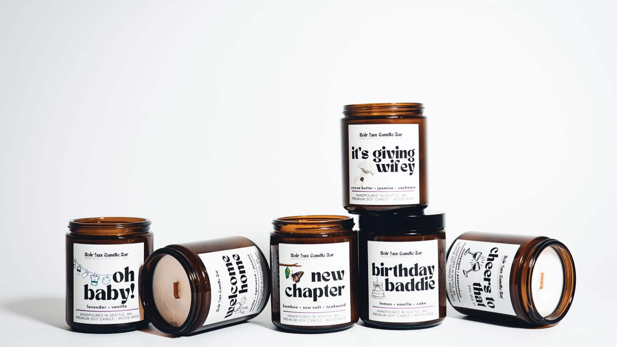 Six amber glass candles with white labels, each featuring different celebratory phrases, are arranged on a plain white background. Each Noir Lux candle exudes a unique fragrance, adding an extra touch of elegance to any occasion.