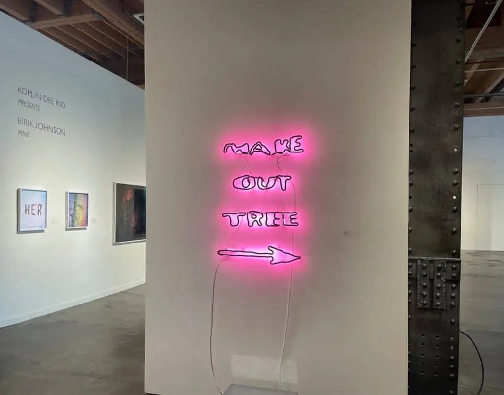 A neon pink sign reading "MAKE OUT TREE" with an arrow is mounted on a white gallery wall; framed artworks are visible in the background.
