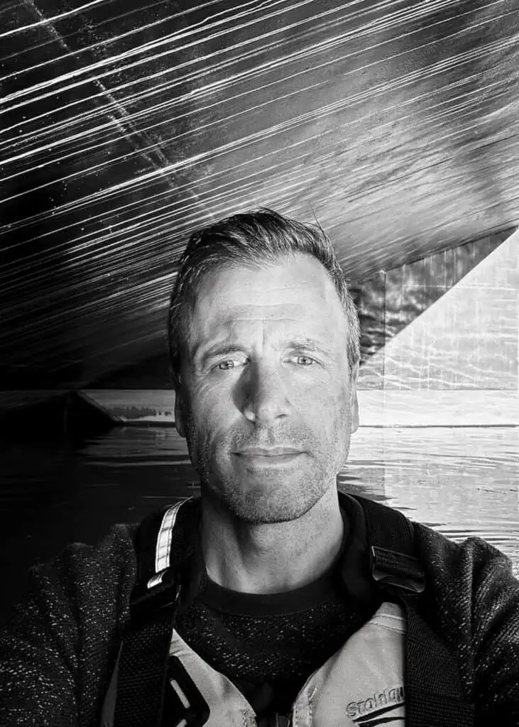 A man wearing a life jacket takes a selfie in front of a reflective surface with linear patterns; water and part of a boat are visible in the background.