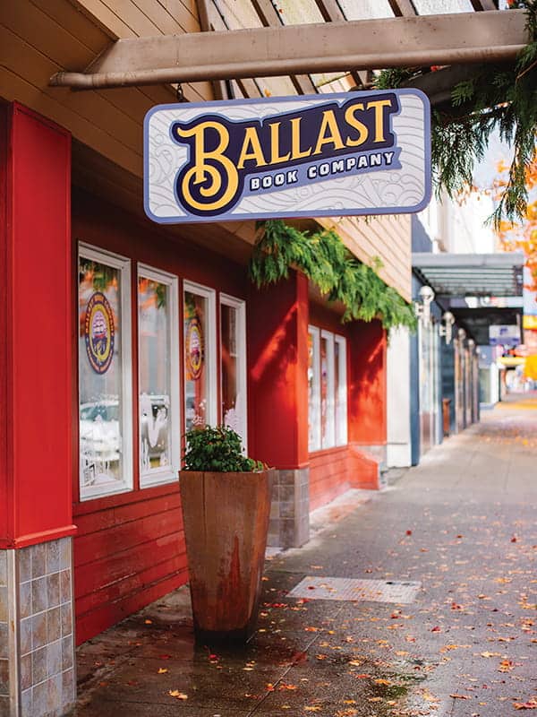A sidewalk view of the exterior of Ballast Book Company, one of Seattle’s beloved independent bookstores, with a large sign above the entrance and autumn leaves scattered on the ground.