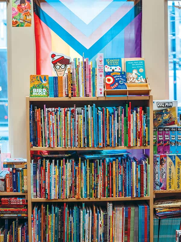 A bookshelf filled with children’s books stands in front of a window displaying a Progress Pride flag, capturing the welcoming spirit of Bainbridge Island—perfect for a summer day trip after a scenic Puget Sound ferry ride.