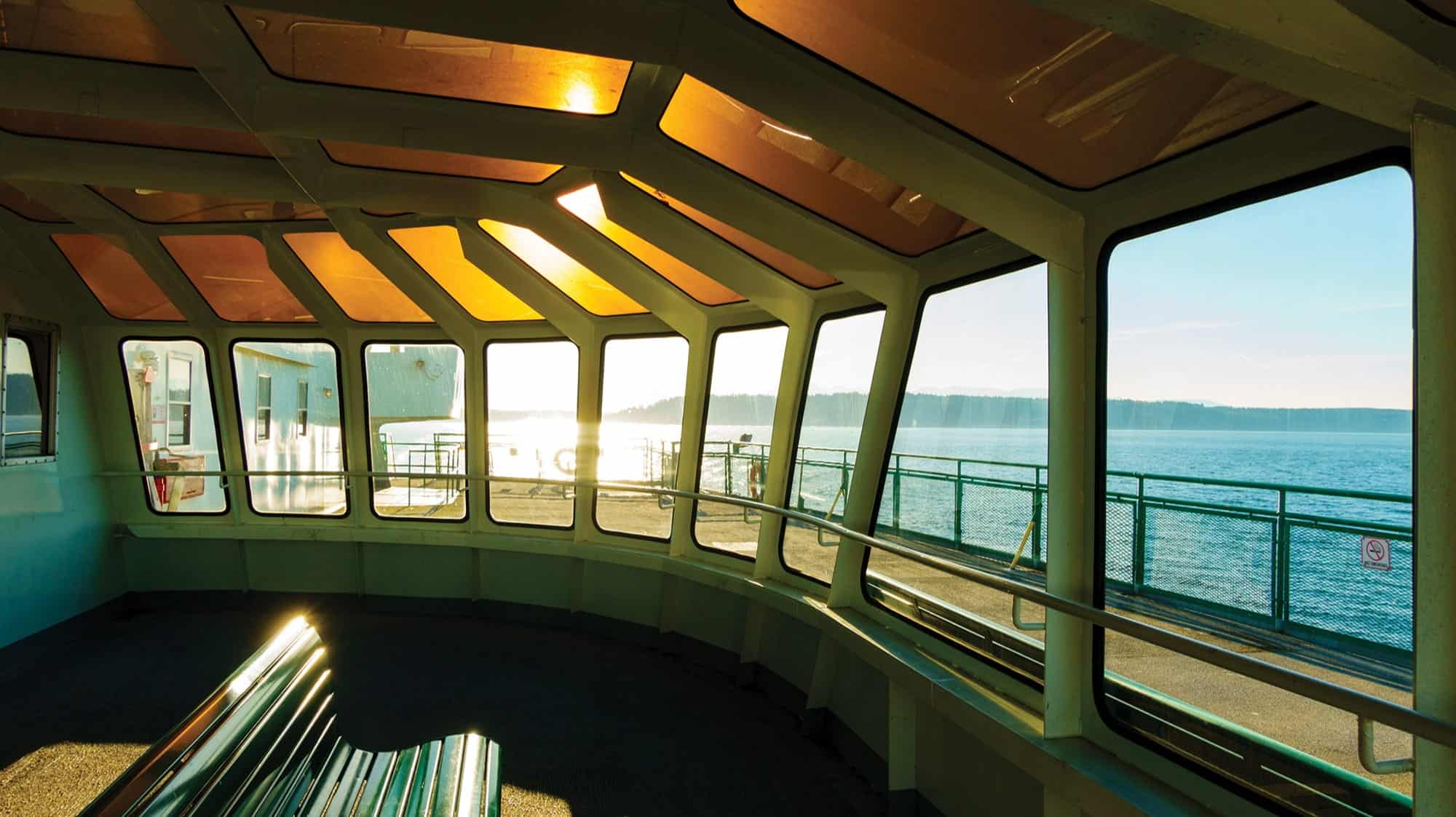 Interior view of a ferry on a summer day trip from Seattle to Bainbridge Island, with large windows, empty benches, and sunlight streaming in, revealing water and the distant shoreline outside.