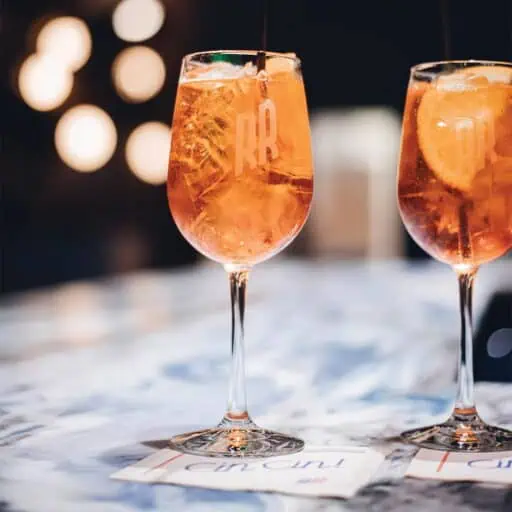 Two wine glasses filled with orange-colored cocktails with ice and orange slices sit on a marble surface next to paper coasters, capturing the vibrant spirit of Capitol Hill's favorite eat and drink dining spots.