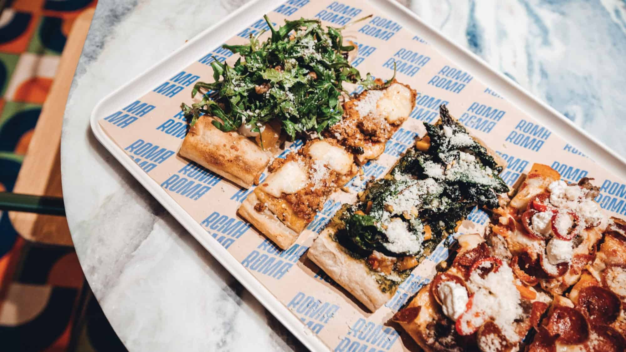 A tray with four rectangular slices of pizza, each topped with different ingredients including arugula, spinach, pepperoni, and cheese, on Roma-branded paper—a delicious example of what you might find at new Seattle restaurants and Capitol Hill dining spots.