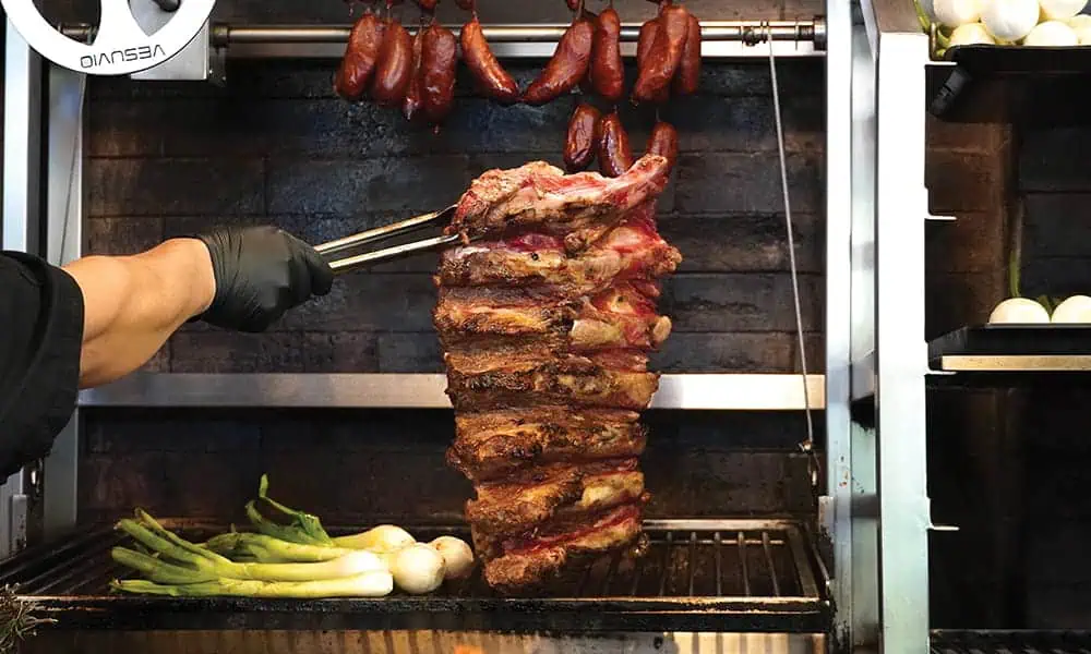 A person uses tongs to grill stacked ribs with green onions; smoked sausages hang above on a rack in a restaurant kitchen—one of the standout dining spots near Capitol Hill.