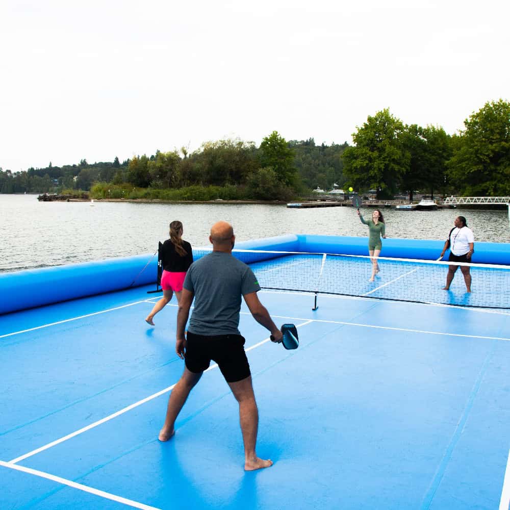 Four people play pickleball on a bright blue floating court on a lake, surrounded by trees and a cloudy sky—an unforgettable scene perfect for any Father’s Day gift guide.