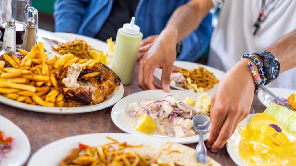 A table with plates of fries, roasted chicken, salad, pasta, and other assorted dishes as two people serve themselves food.