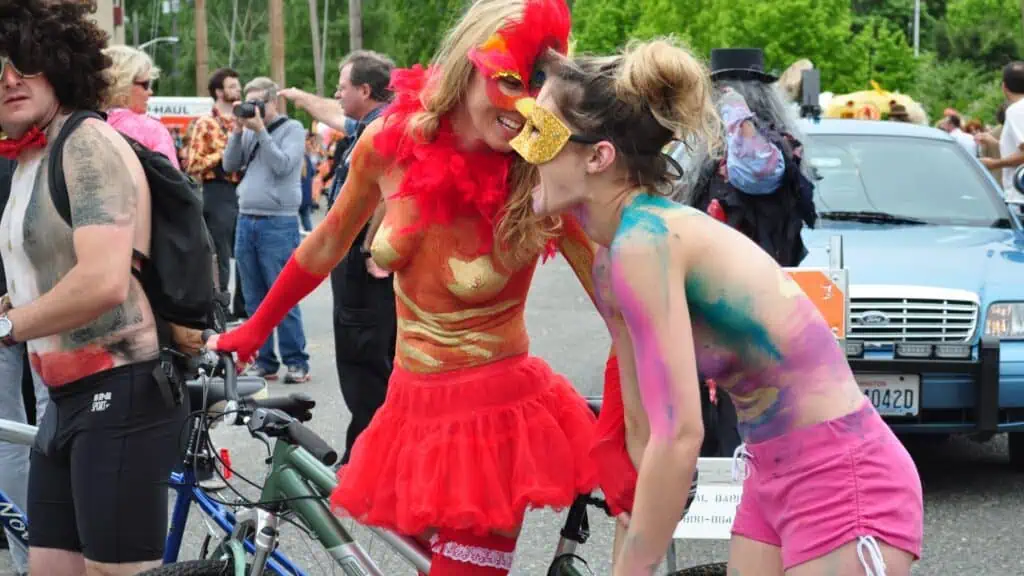 Two people in colorful body paint and costumes stand next to bicycles at an outdoor event, surrounded by other participants and parked cars.