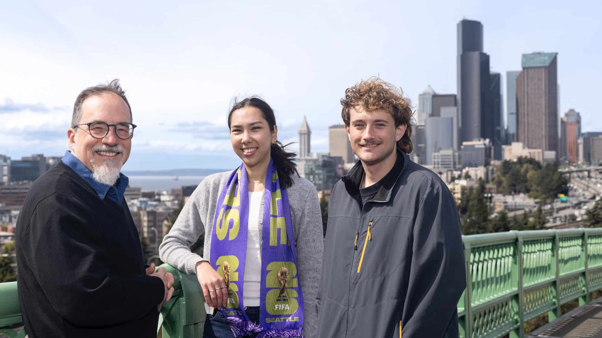 Three people stand on a bridge overlooking the Seattle skyline with tall buildings in the background on a clear day.