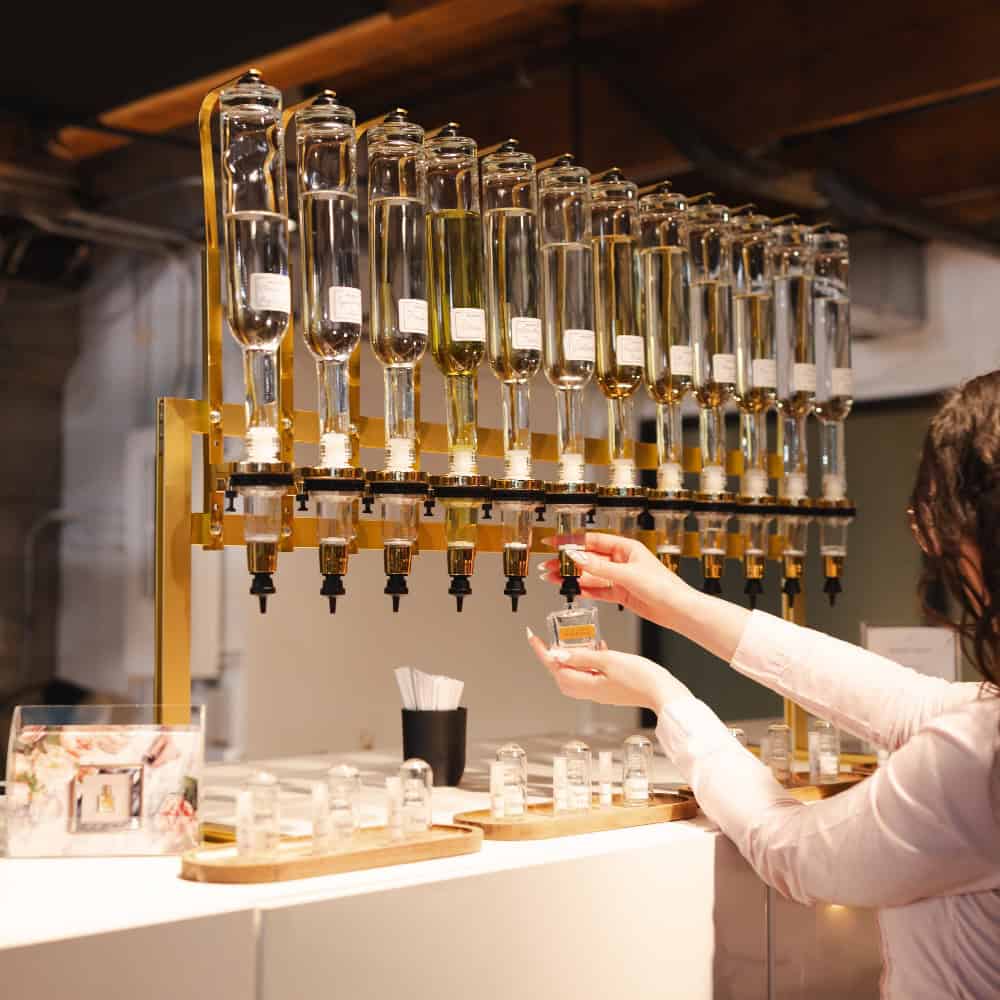 In a well-lit indoor setting reminiscent of a classic parfumerie, a person fills a small glass bottle from a row of large glass dispensers containing various liquids.