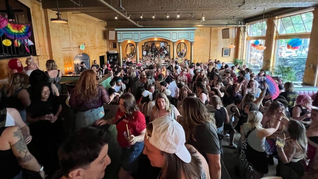 Large crowd of people socializing inside a bar with wooden walls and colorful decorations, with natural light coming through big windows.