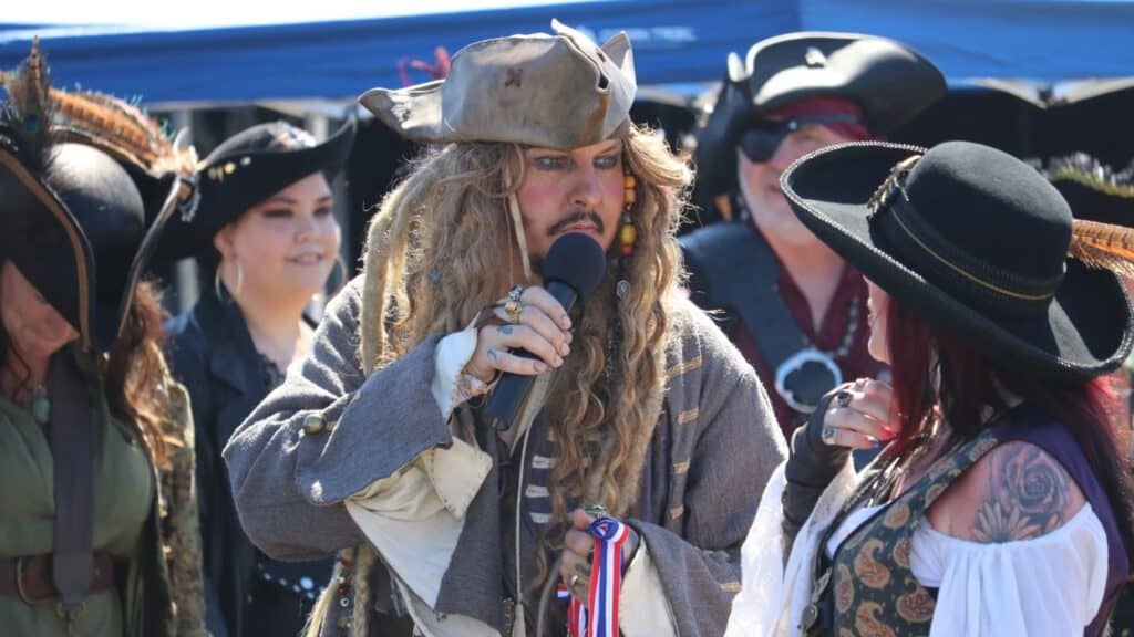 At a lively Washington summer festival, a person dressed as a pirate speaks into a microphone, surrounded by others in weird and wonderful pirate costumes.
