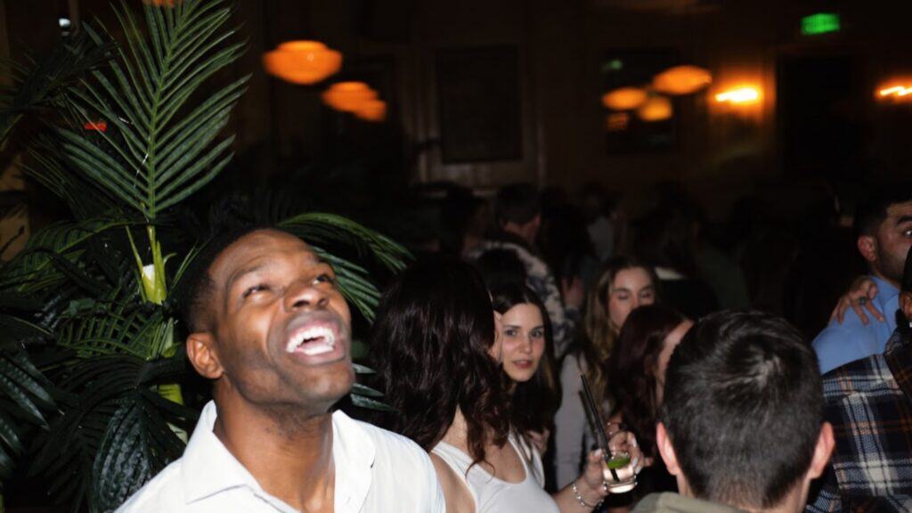 A man in a white shirt smiles broadly in the foreground of a dimly lit, crowded indoor party with plants and people in the background.