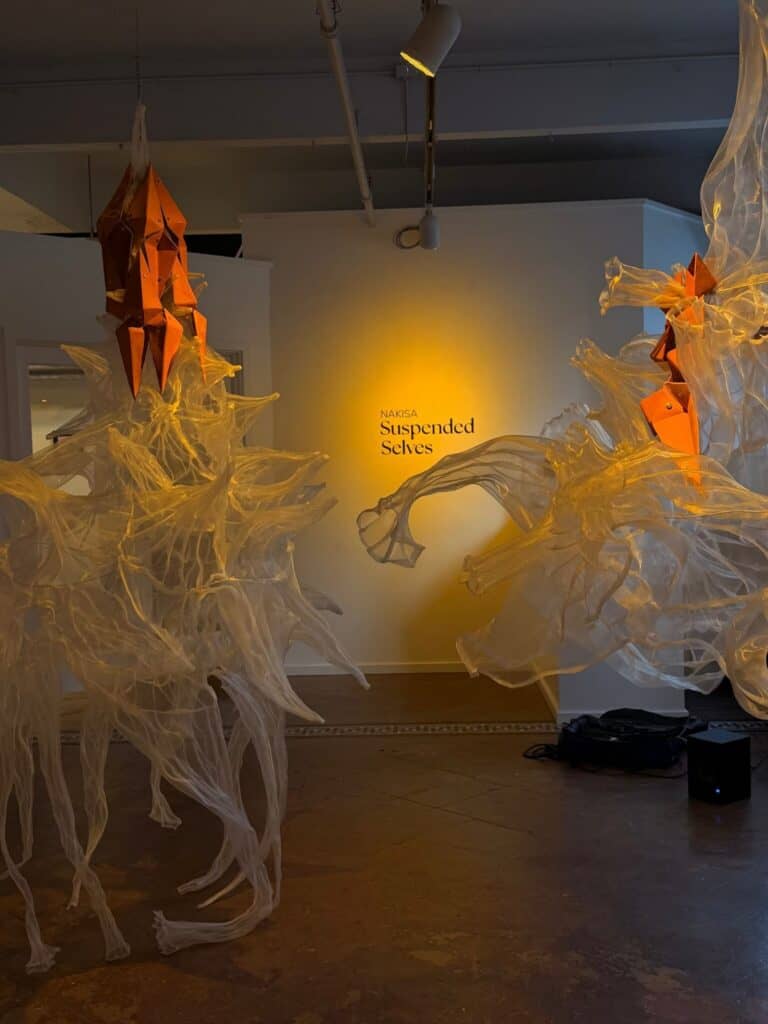 Art installation featuring translucent, flowing fabric structures suspended from the ceiling with orange geometric forms, displayed in a gallery with "Suspended Selves" text on the wall.
