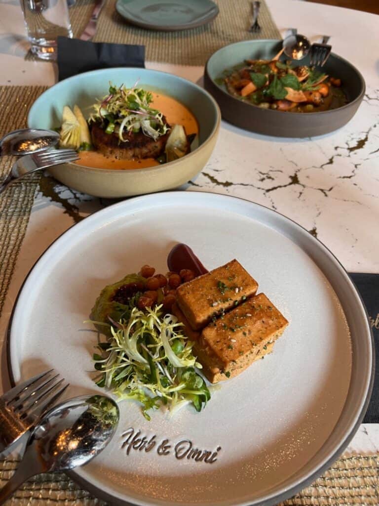 Three plated dishes are arranged on a table, each featuring plant-based meals with greens and garnishes at a restaurant called Herb & Omni.