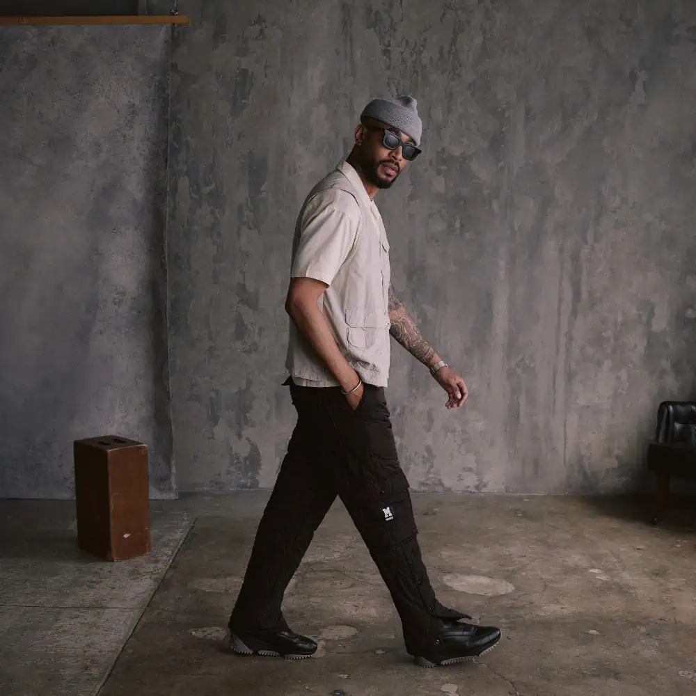 A man in a light shirt, black pants, beanie, and sunglasses walks indoors on a concrete floor with a textured gray wall—perfect inspiration for your Father’s Day Gift Guide.
