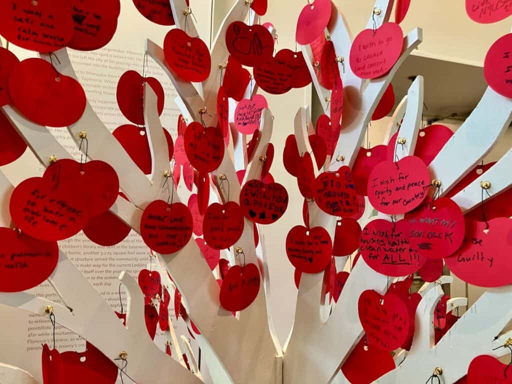 A white tree sculpture decorated with red paper apple cutouts, each containing handwritten messages and drawings.