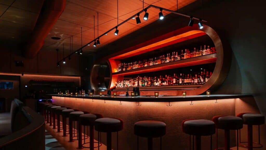 Modern bar interior with a curved illuminated bar counter, high stools, and shelves stocked with bottles against a warm, ambient lighting backdrop.