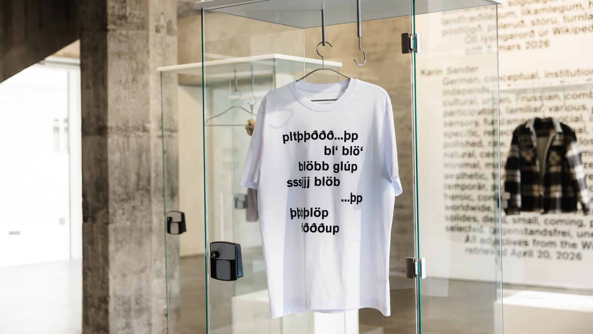 A white t-shirt with black abstract text hangs inside a glass display case in a modern gallery space.