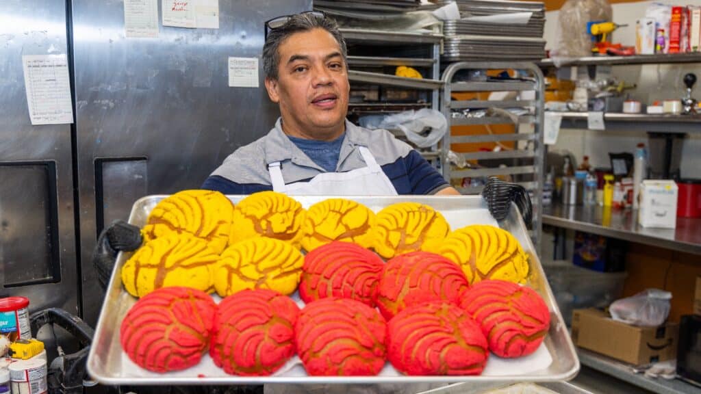A person in a bakery kitchen holds a tray filled with freshly baked yellow and pink conchas, a type of Mexican sweet bread, with baking supplies visible in the background.