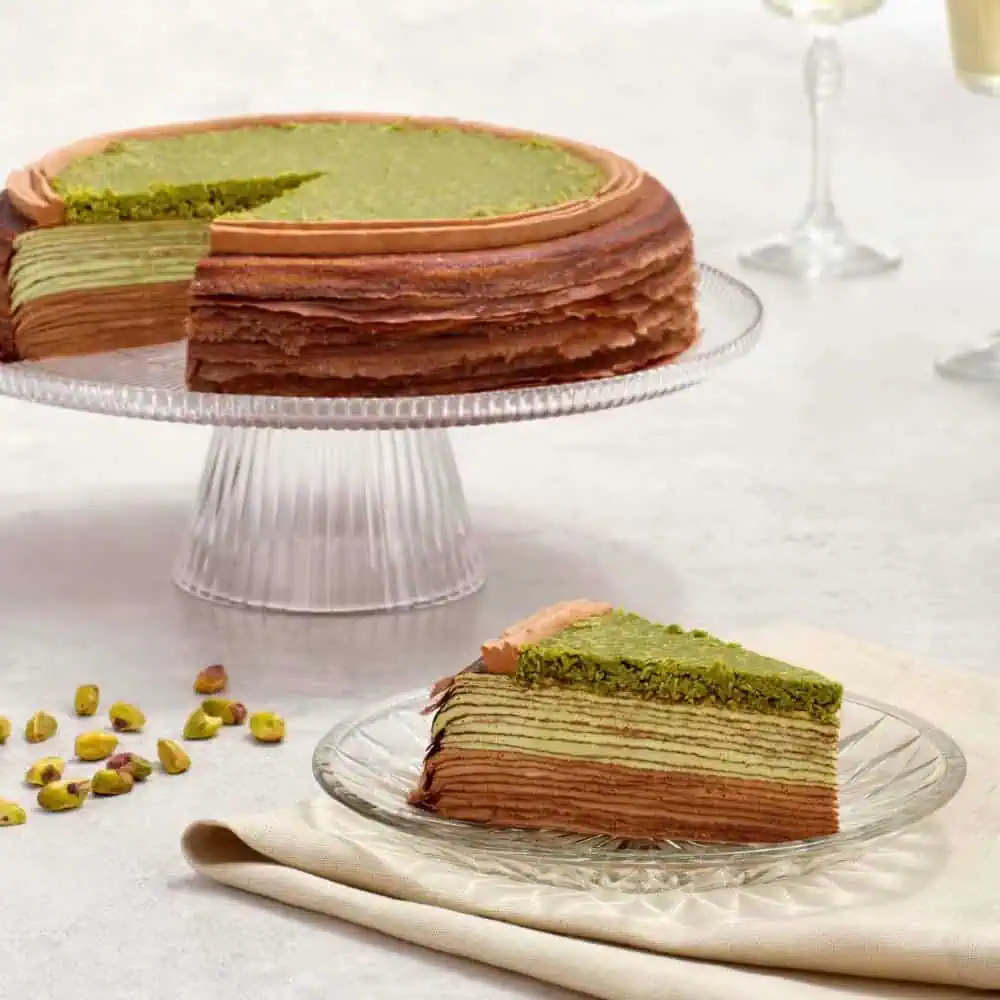 A slice of layered green and brown crepe cake sits on a plate, with the rest of the cake and some pistachios visible on a glass cake stand in the background.