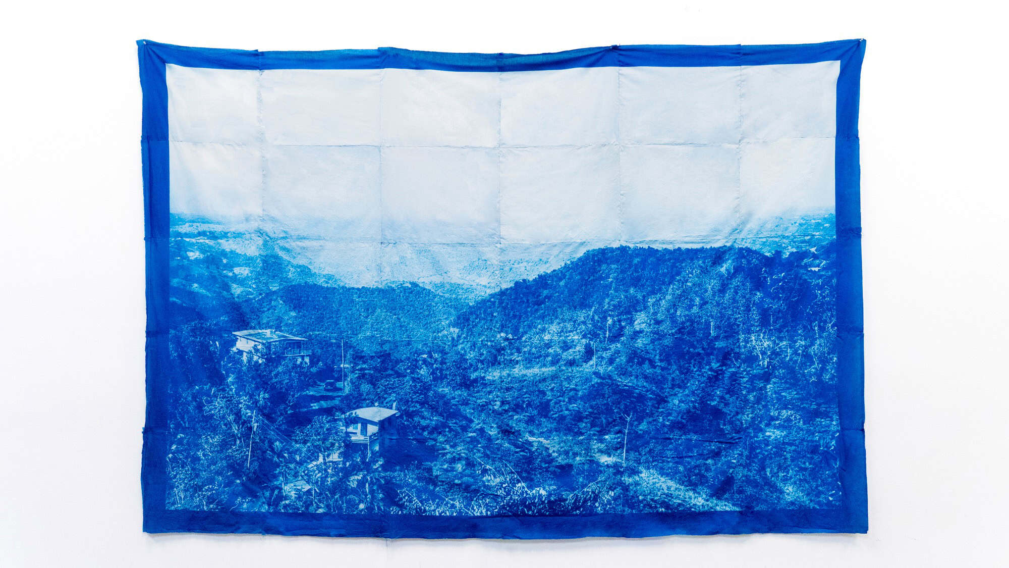 A large blue-toned fabric artwork displays a photographic image of a mountainous landscape with a few houses among trees.