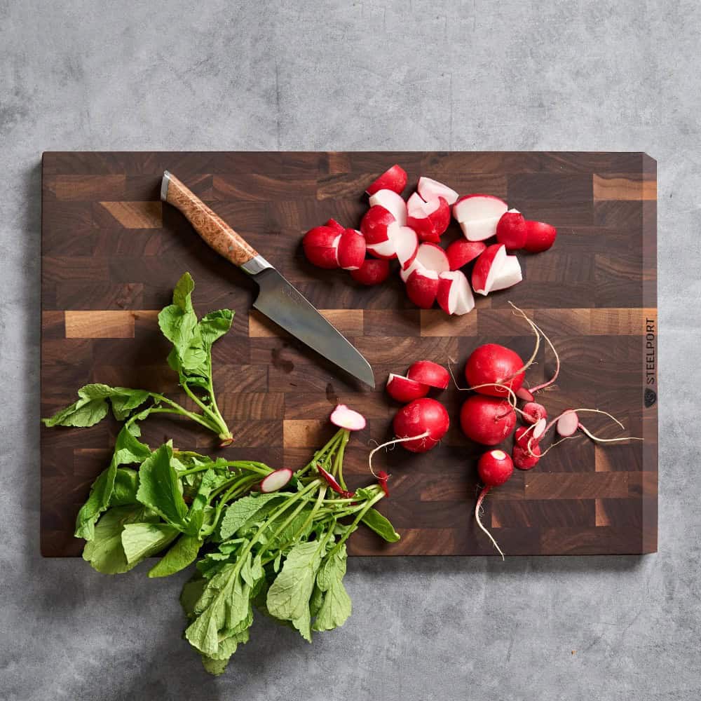 A knife and sliced radishes are on a dark wooden cutting board, with whole radishes and green radish tops on a gray countertop.