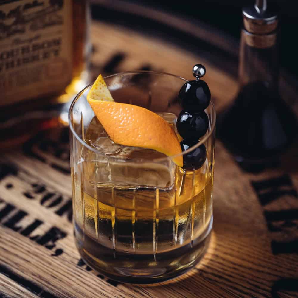 A glass of whiskey with a large ice cube, garnished with an orange peel and dark cherries, sits on a wooden surface next to a bottle and a small glass container.