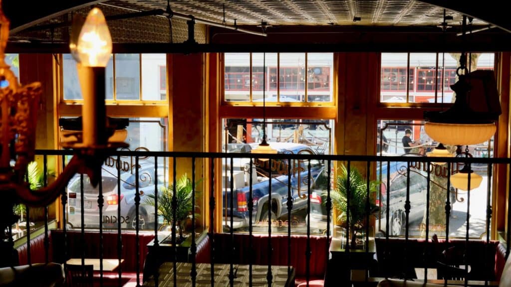 View from inside a restaurant with large front windows, red booths, decorative plants, and parked cars visible outside on a sunny day.