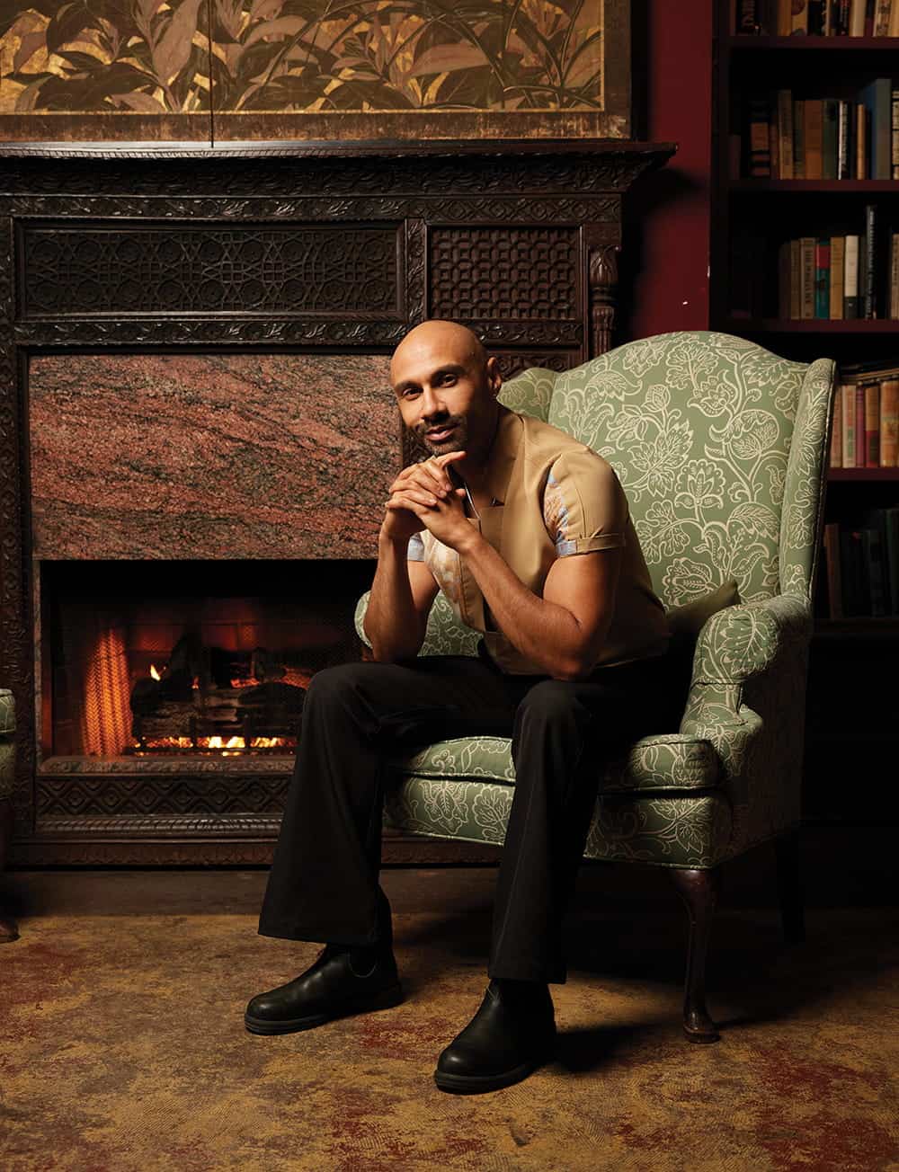 A man sits in a patterned armchair by a fireplace, his hands clasped and elbows on his knees, in a room with bookshelves and ornate decor—capturing the thoughtful ambiance of an Unincarcerated Productions set.