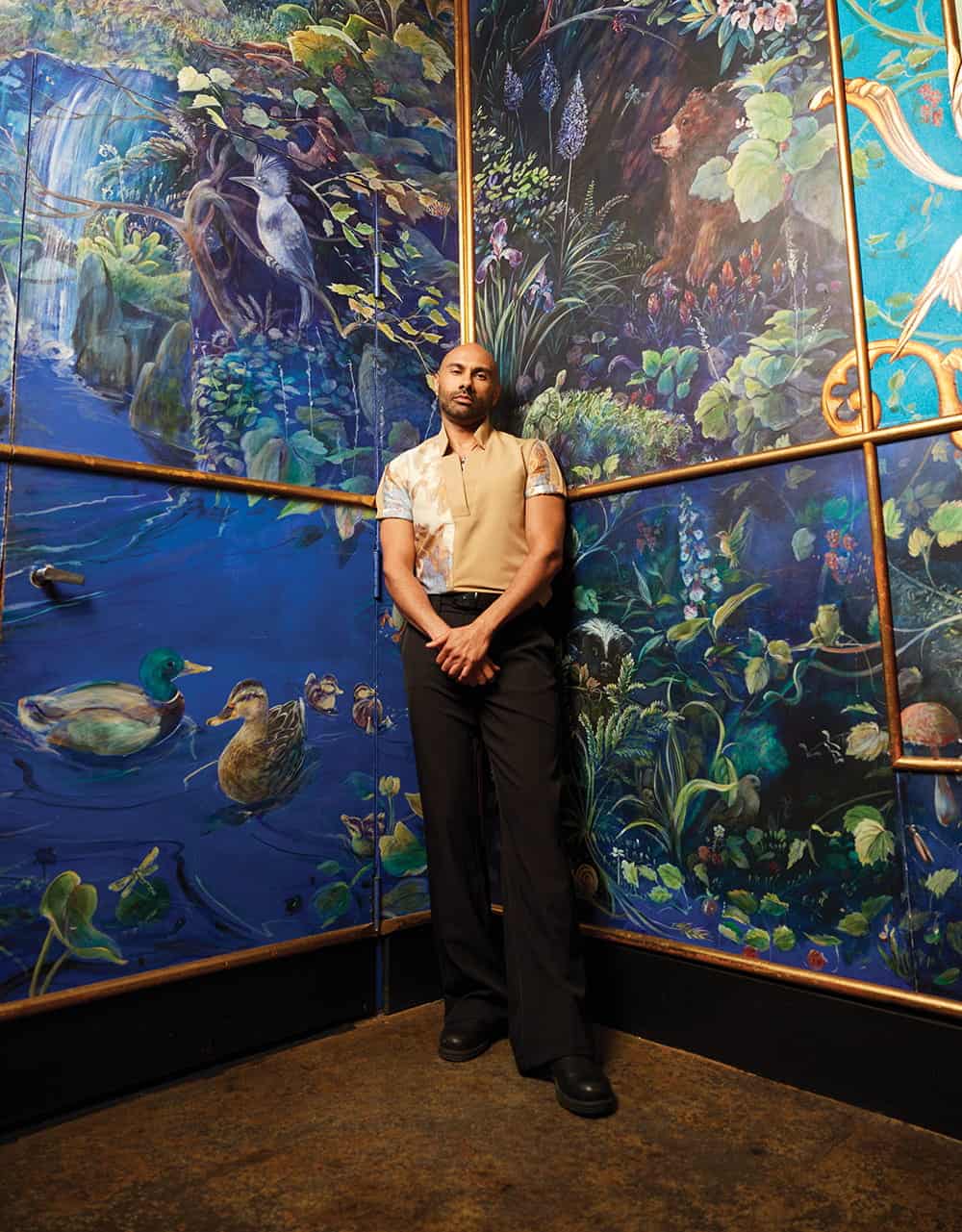 A man stands with hands clasped in front of a vibrant, nature-themed mural featuring birds, ducks, and foliage—a powerful scene from the Washington prison podcast by Unincarcerated Productions.