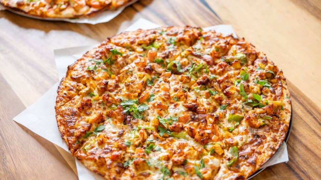 A cooked pizza topped with diced chicken, green bell peppers, herbs, and melted cheese sits on parchment paper on a wooden table.