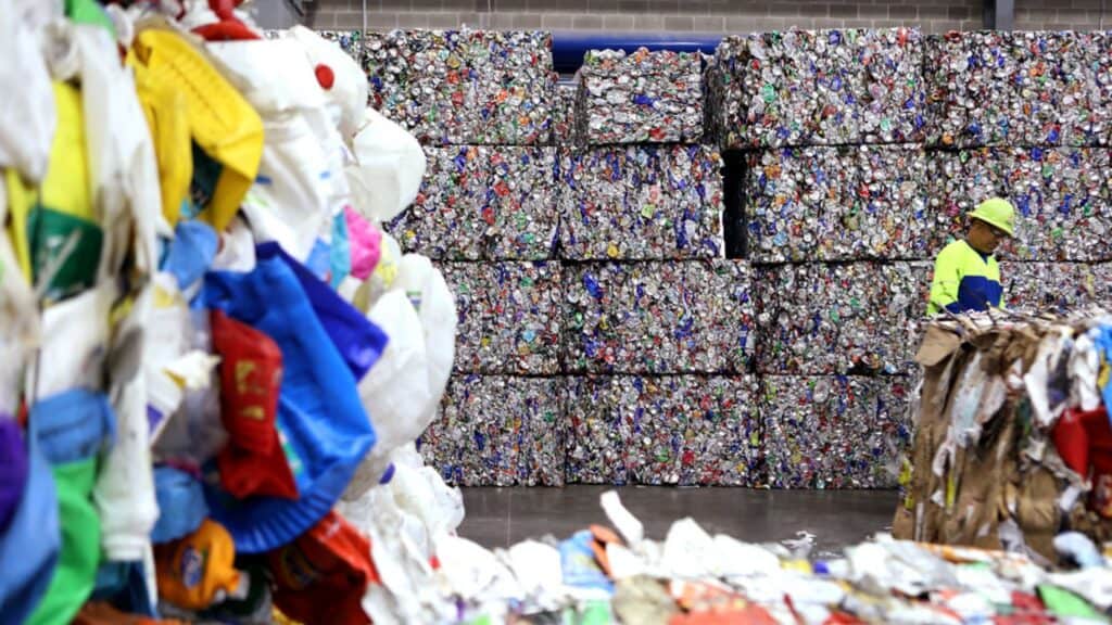 Bales of compacted recyclable materials are stacked in a warehouse, while a worker in a safety vest and helmet inspects the area.