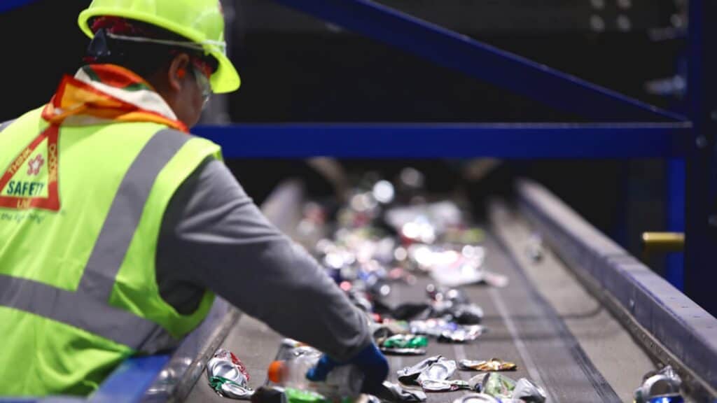 Worker in a safety vest and helmet sorts crushed cans on a conveyor belt at a recycling facility.