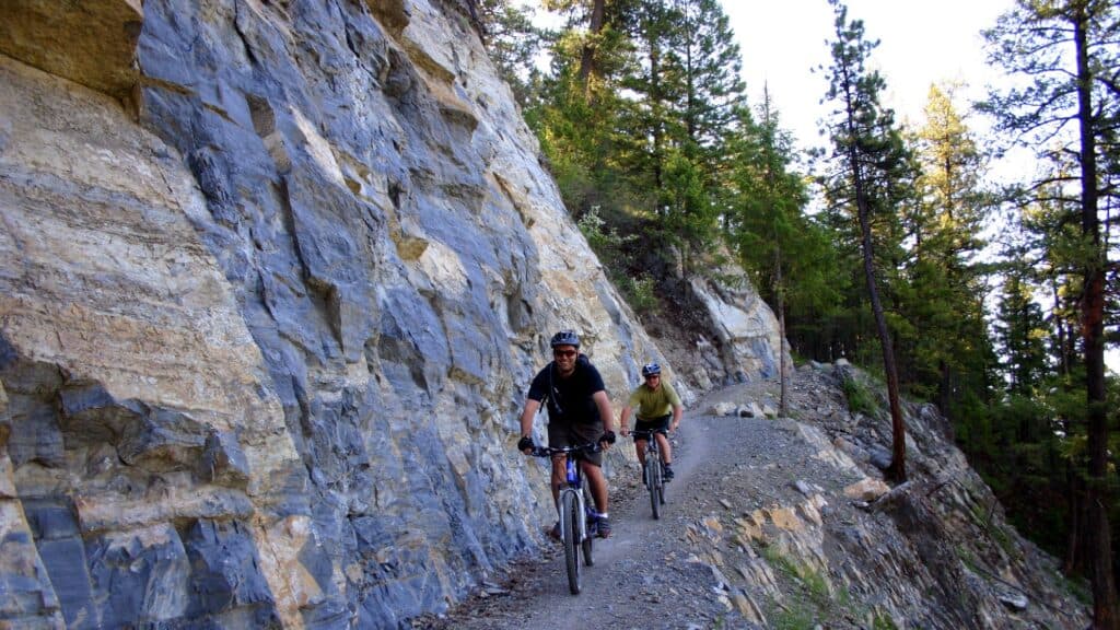 Two people ride mountain bikes on a narrow dirt trail along a rocky cliffside, surrounded by tall trees and forested landscape.