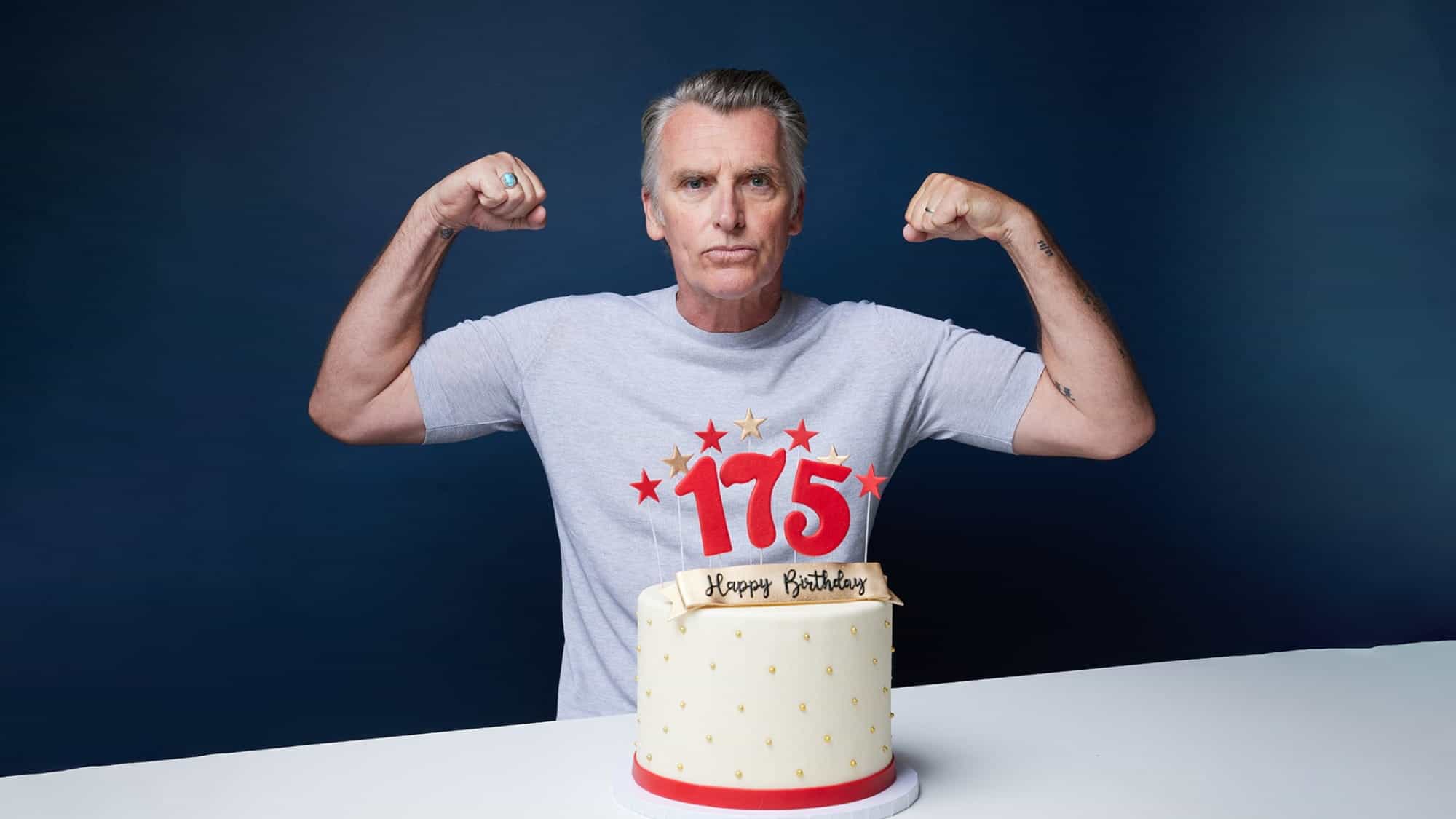 A man in a gray t-shirt, channeling the style of John Pearson, the world's first male supermodel, flexes his arms behind a white birthday cake with red "175" candles and a "Happy Birthday" sign on top.
