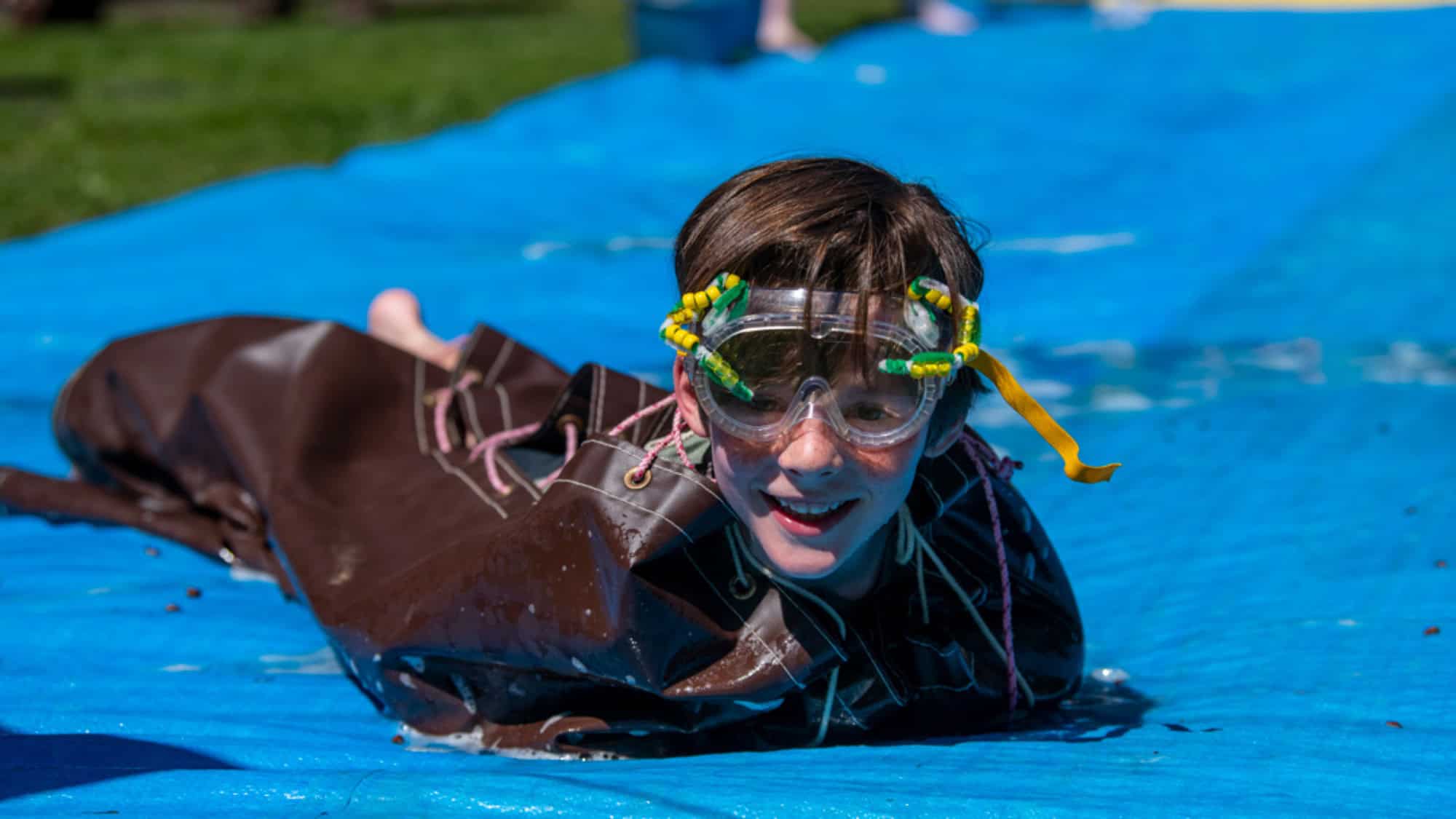 A child wearing goggles and a brown poncho slides on a blue tarp outdoors, smiling and looking forward.