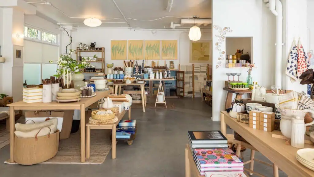 A well-lit boutique store interior with wooden tables displaying home decor, ceramics, books, and various household items, against white walls and concrete floors.