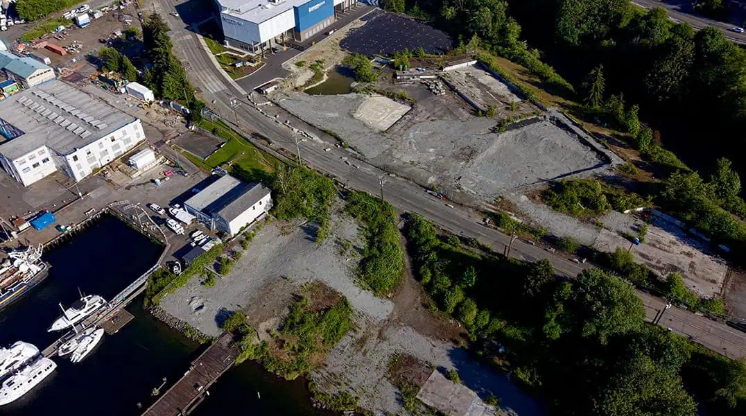Aerial view of an industrial area with vacant lots, several buildings, parked boats at a dock, and a road running through the center—where exotic automobiles reminiscent of Formula One lineups could cruise, bordered by trees and greenery.