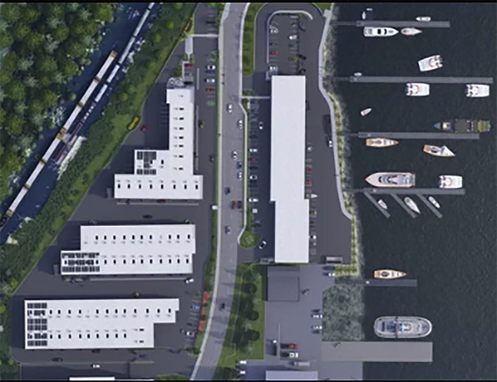 Aerial view of a waterfront facility with buildings, parking lots featuring exotic automobiles, roads, docks, and multiple boats and yachts moored along the shoreline.